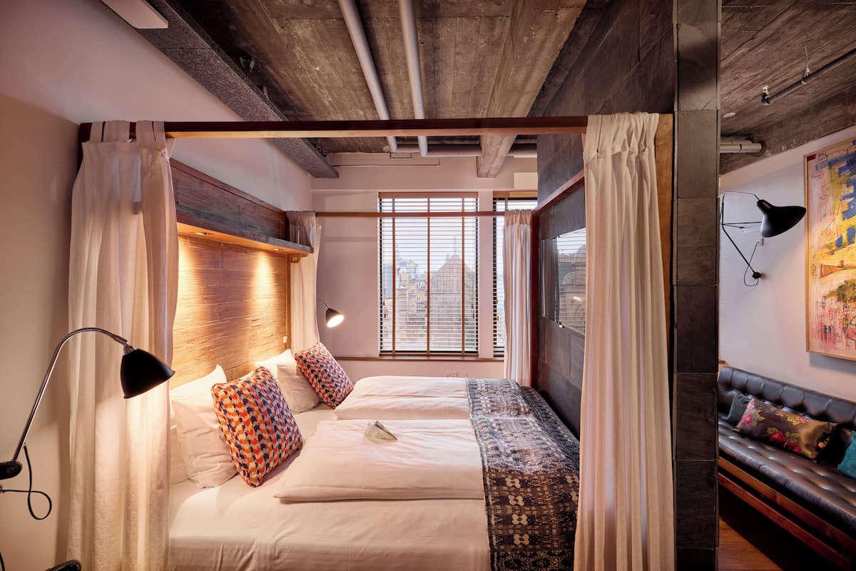 Ein modernes Hotelzimmer mit einem großen Bett, einer Couch und Blick auf die Stadt durch große Fenster.