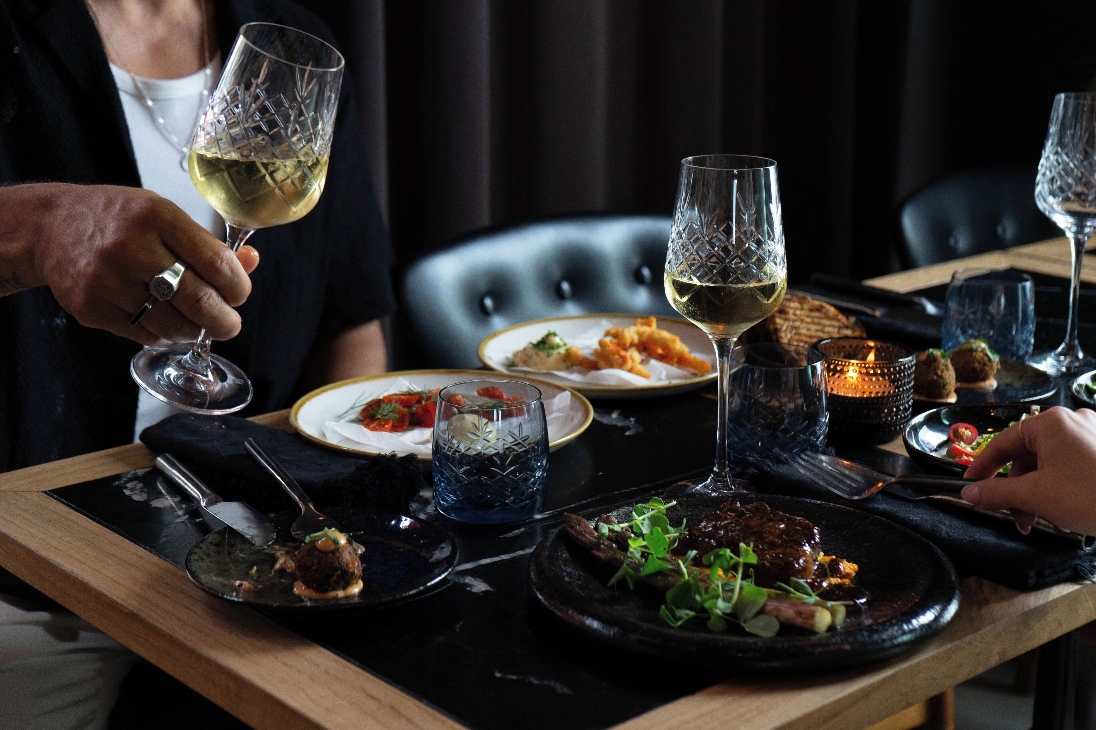 Una mesa de comedor con varios platos y copas de vino, ubicada en una sala con poca luz y una silla de cuero negro al fondo.