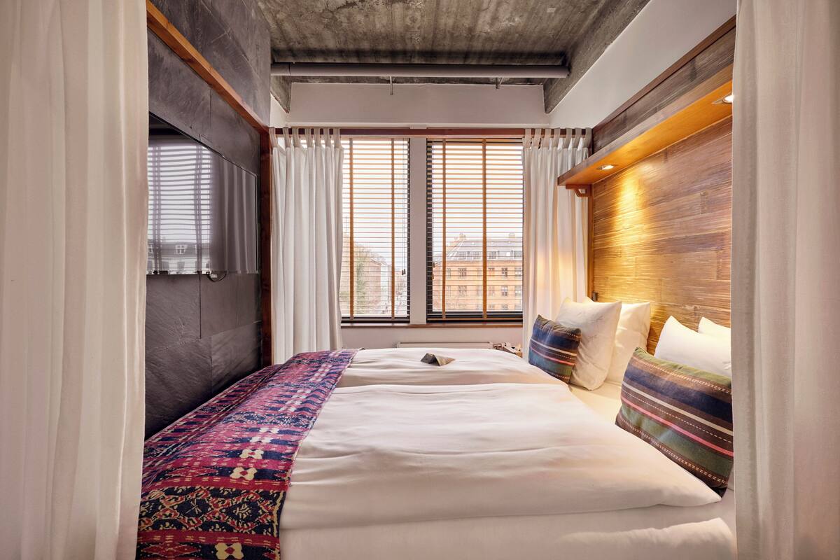 A modern hotel room with a large bed, a wooden headboard, and a view of the city through floor-to-ceiling windows.