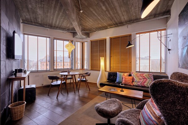Una sala de estar moderna con piso de madera, un sofá con cojines de colores, una mesa de comedor con sillas y ventanas grandes con persianas.