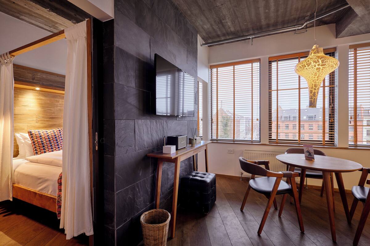 Une chambre d’hôtel moderne équipée d’un lit, d’un téléviseur fixé au mur, d’une table à manger avec des chaises et de grandes fenêtres avec des stores.