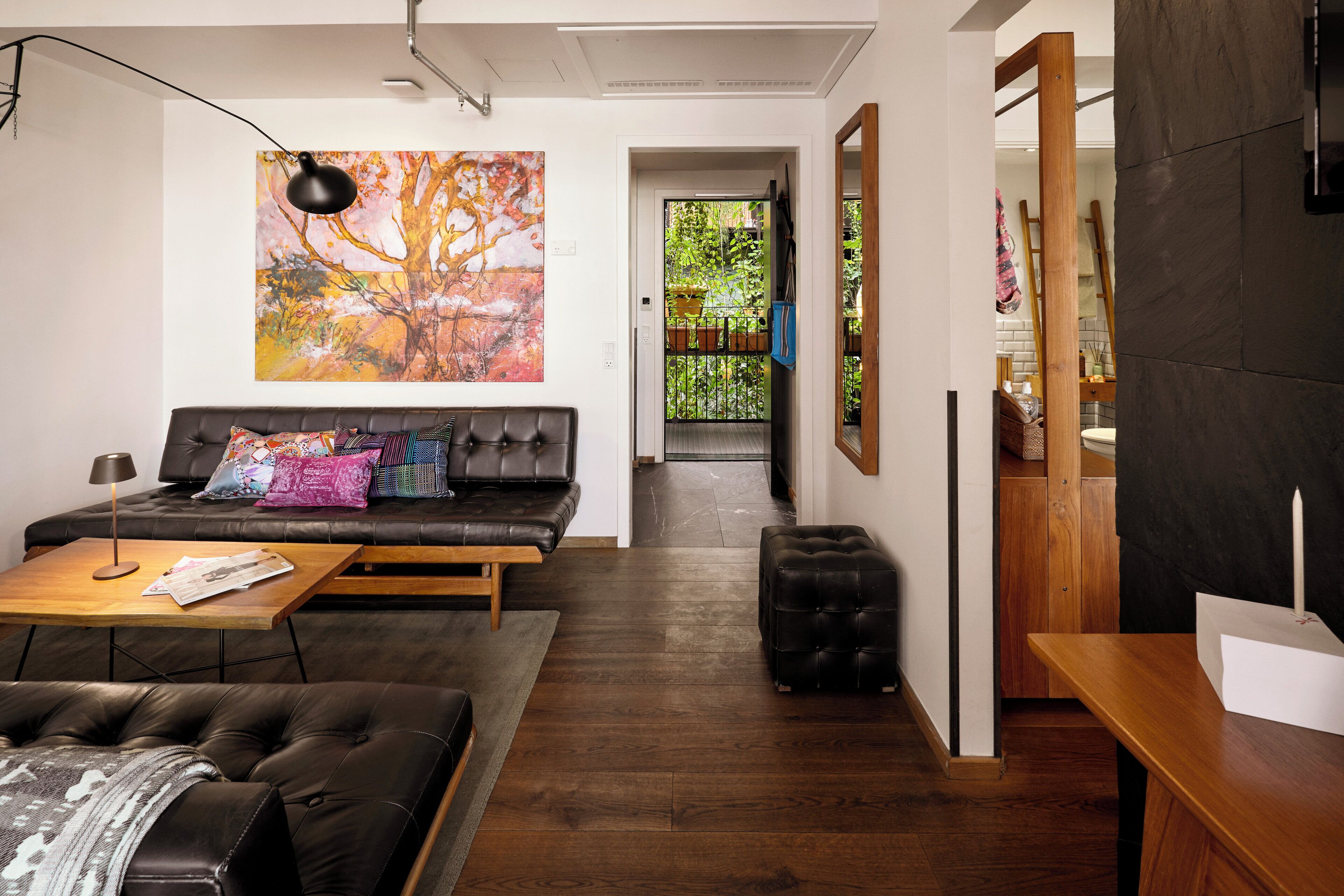 A modern living room with a sofa, coffee table, and ottoman, a painting on the wall, and a view into the kitchen area.