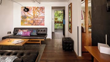 A modern living room with a sofa, coffee table, and ottoman, a painting on the wall, and a view into the kitchen area.