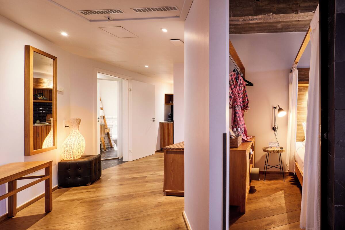Ein Hotelzimmer mit Holzboden, einer Bank, einem Nachttisch, einer Lampe und einem Bett mit weißem Vorhang.