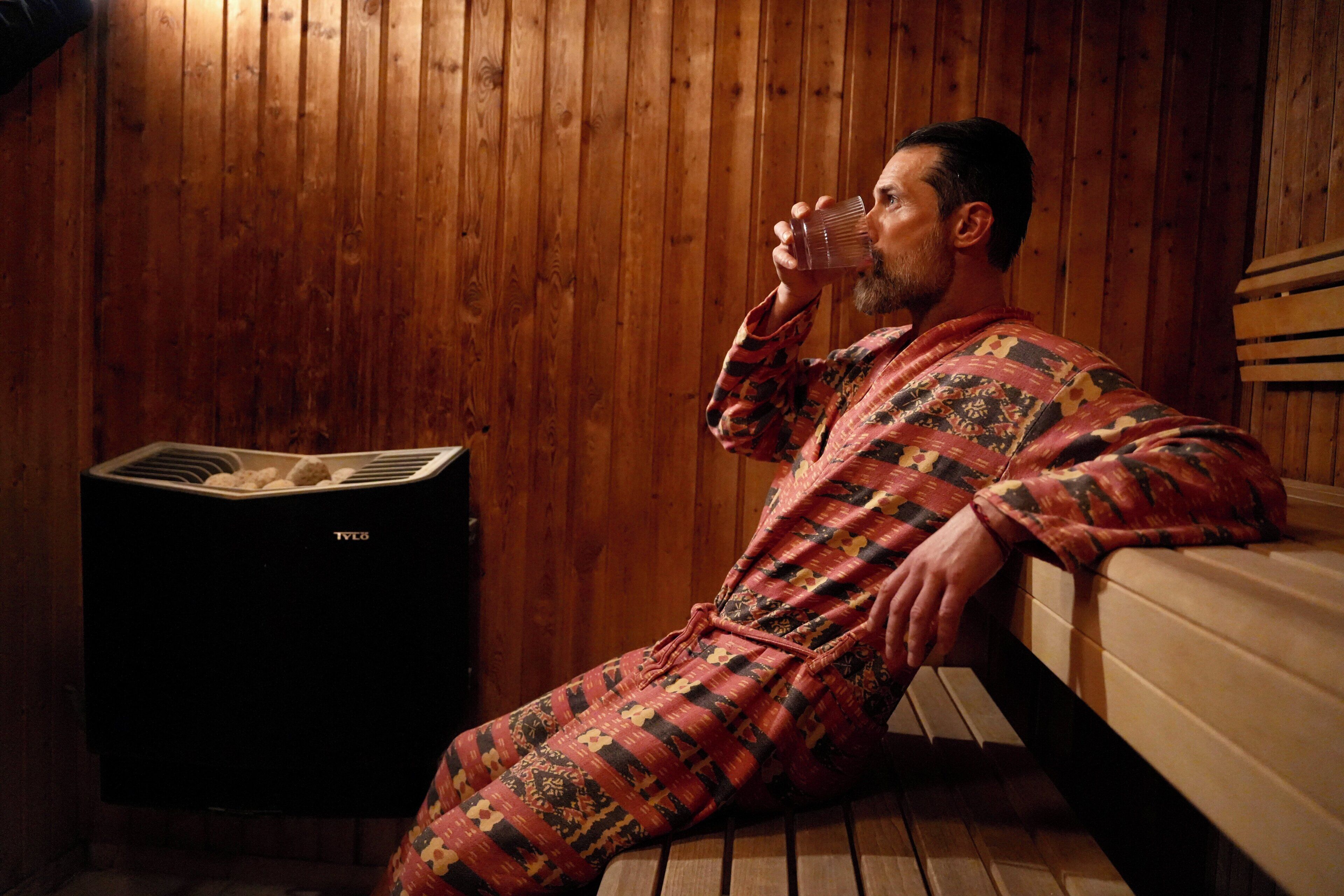 Une personne vêtue d’une robe à motifs est assise sur un banc dans une pièce aux murs en bois, qui contient également un sauna.