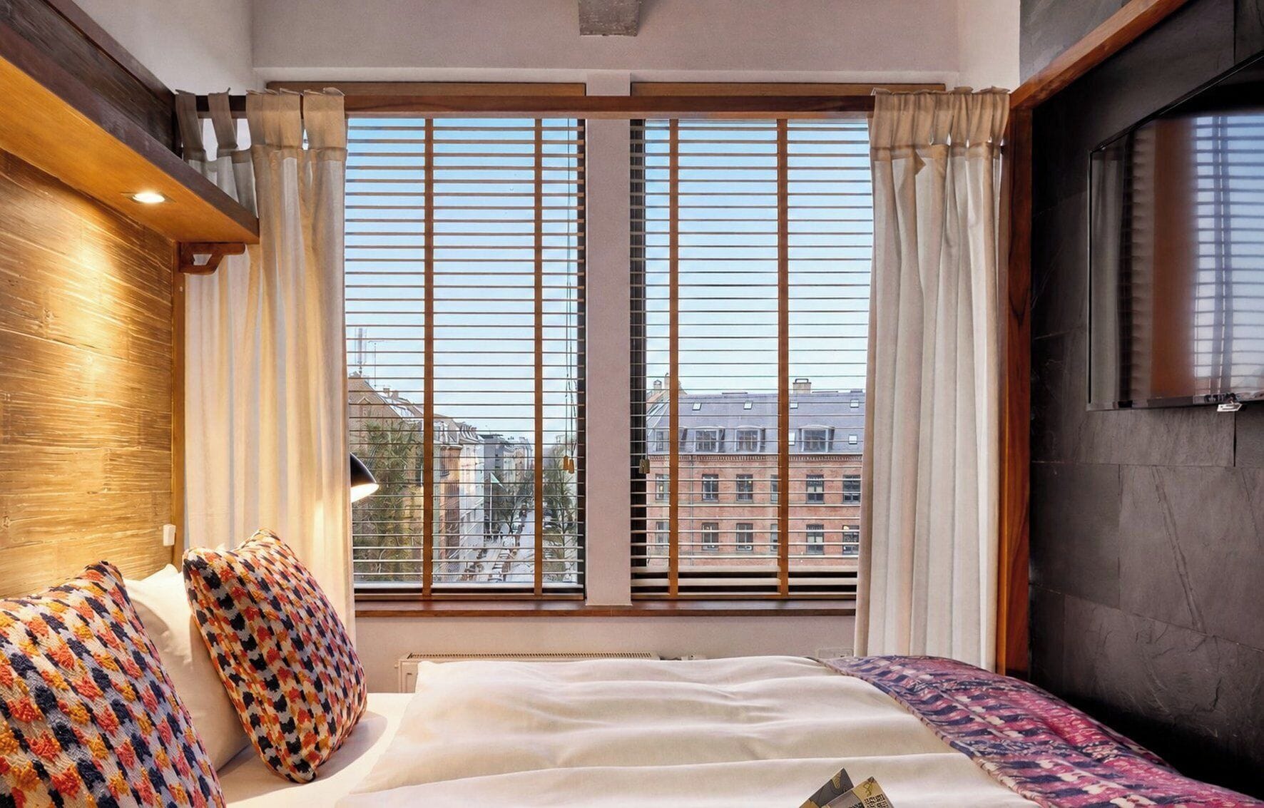 A hotel room with a large window, a bed with patterned pillows, and a view of buildings.
