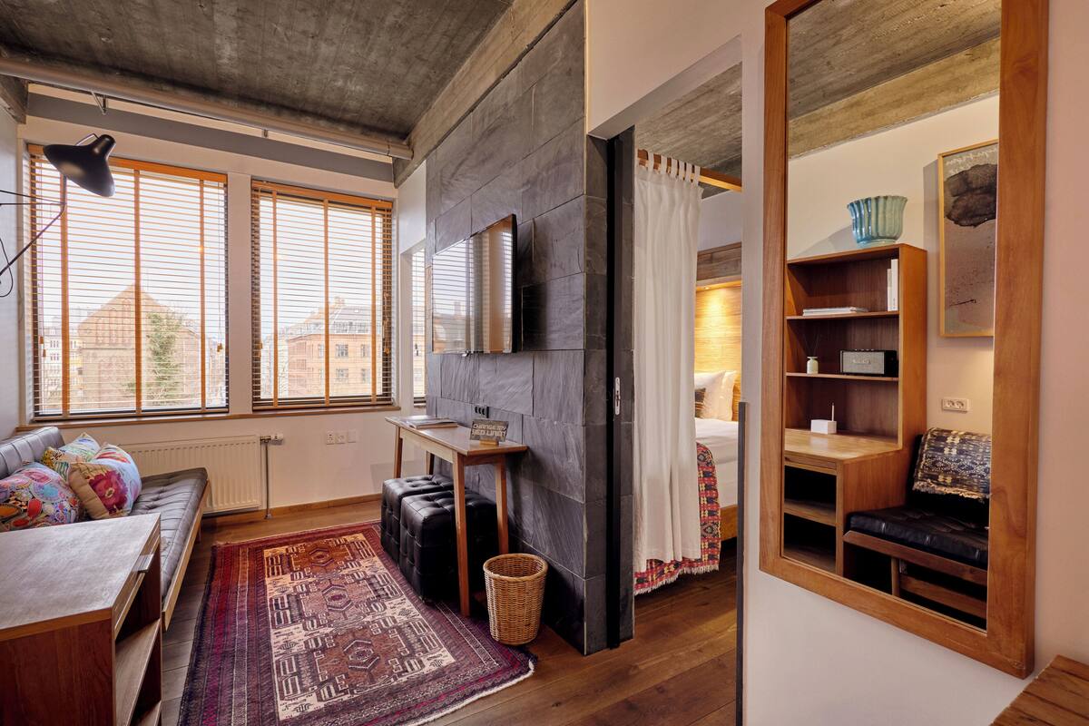 Un salon moderne avec un canapé, un pouf et une grande fenêtre équipée de stores.