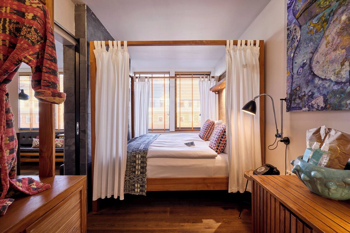 A bedroom with a wooden bed, white curtains, a bedside lamp, a large window, and a painting on the wall.