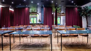 Un salón de conferencias con mesas y sillas, ventanas amplias con cortinas y una planta decorativa.