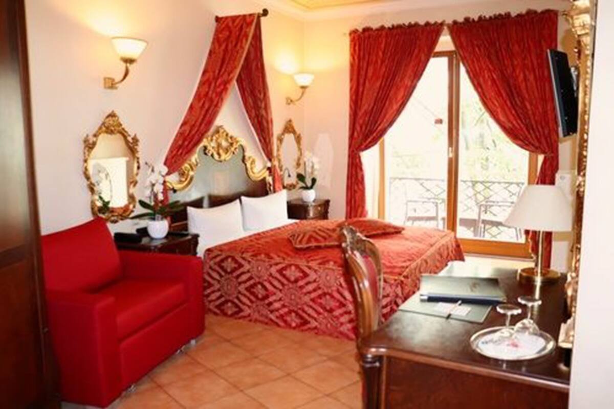 Een hotelkamer met een bed, een rode bank, een bureau met lamp en uitzicht op een balkon.