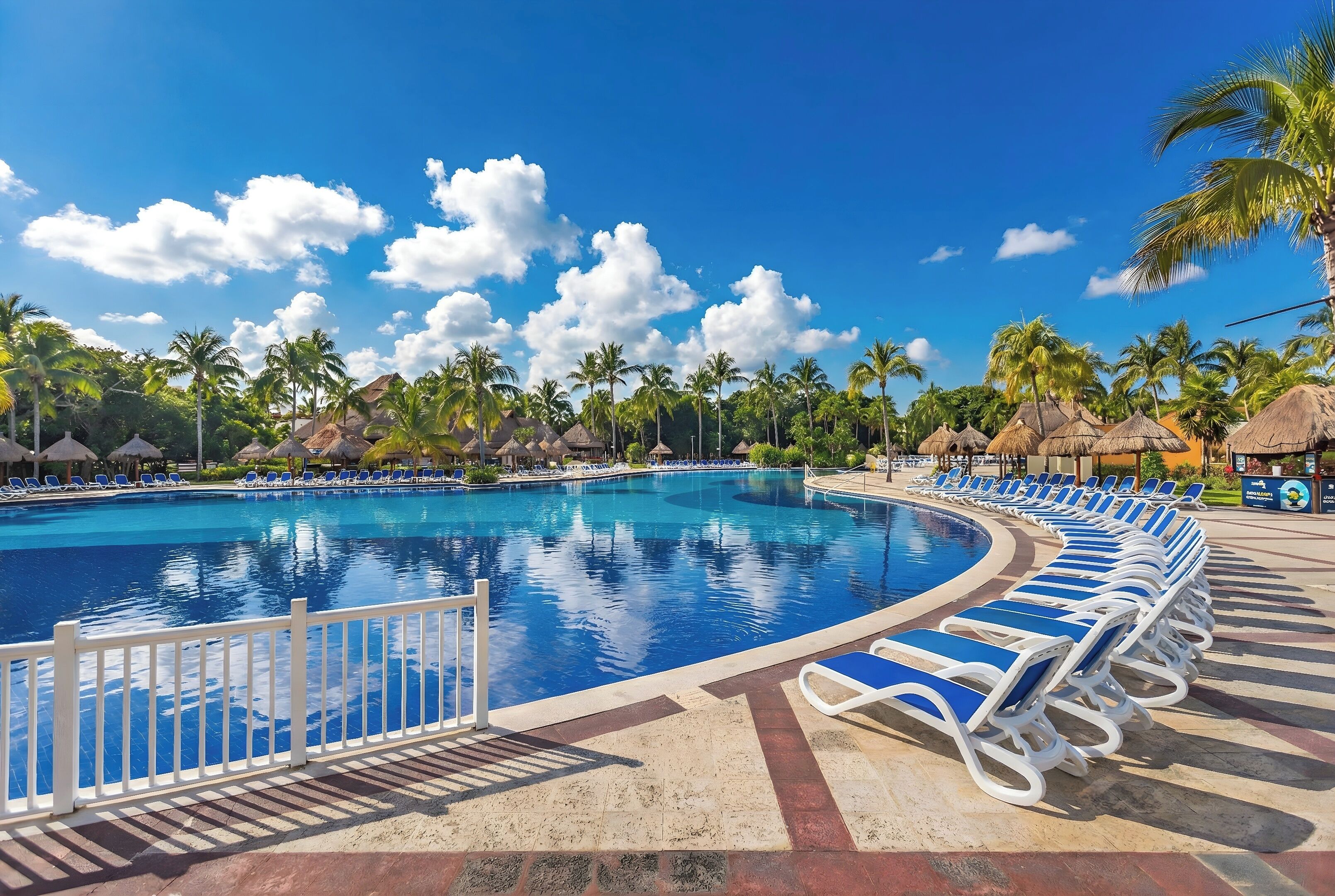 Una piscina con sillas de descanso, palmeras y estructuras de techo de paja.