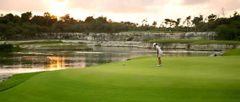A golf course with a water hazard, a golfer in mid-swing, and a sunset in the background.