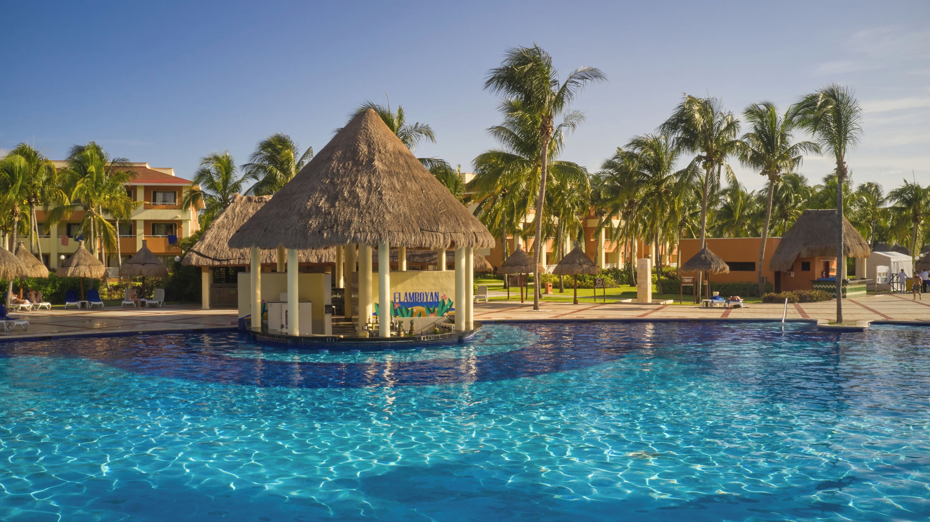 Una piscina con una estructura de techo de paja, palmeras y un edificio con la señalización 'ELAMBROYAN'.