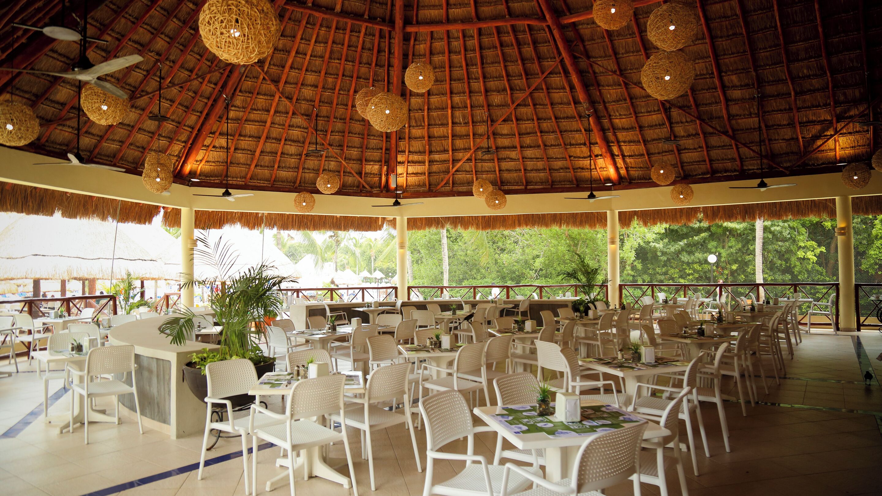 A spacious indoor area with thatched ceilings, white plastic chairs, and tables, featuring large windows and a view of greenery outside.