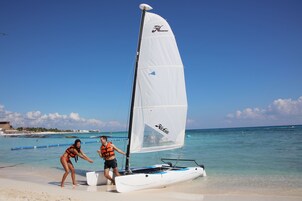 Escena de playa con un velero y dos personas cerca del agua.