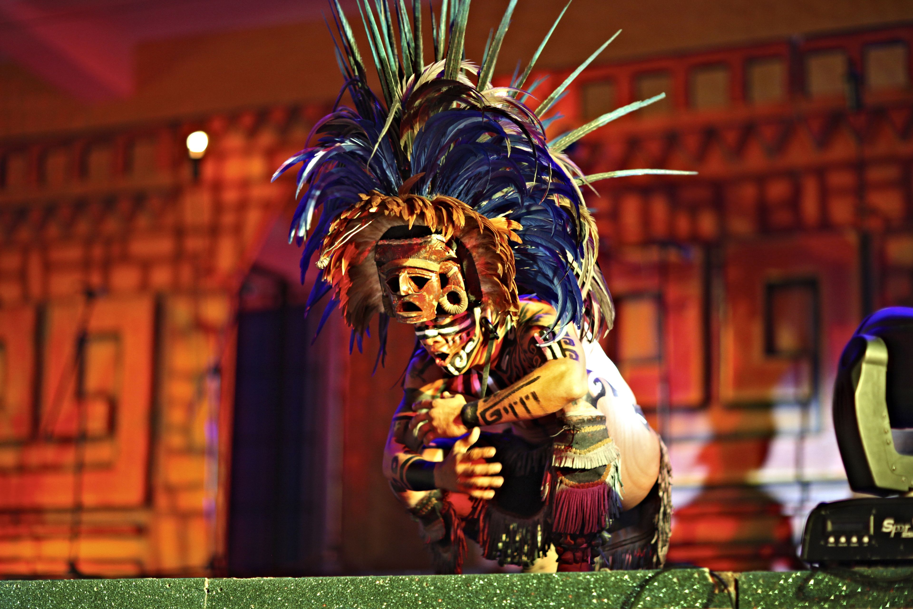 A person in traditional attire with a feathered headdress, standing in front of a building with a red facade.