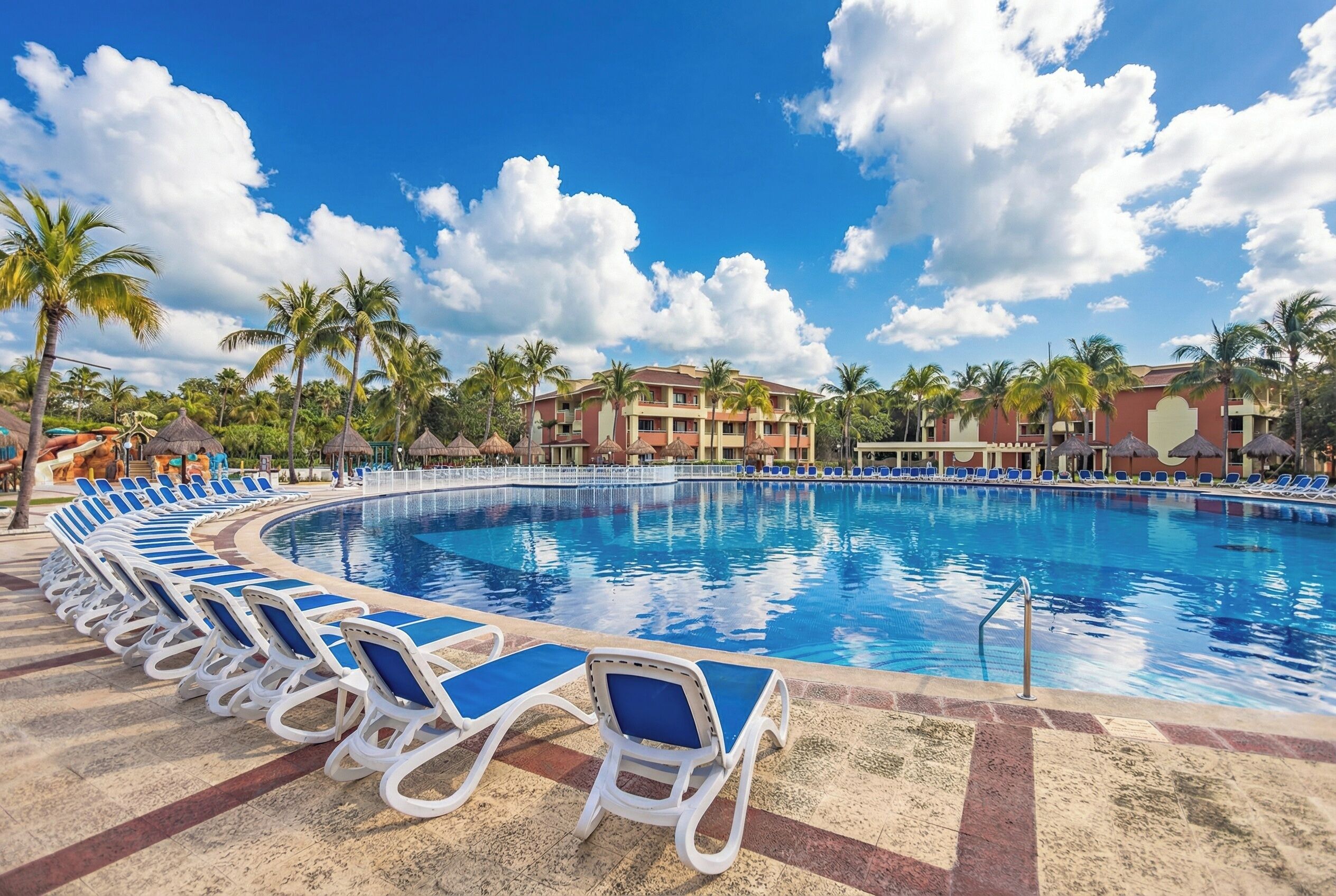 A swimming pool with lounge chairs, palm trees, and a building with a red and beige facade.