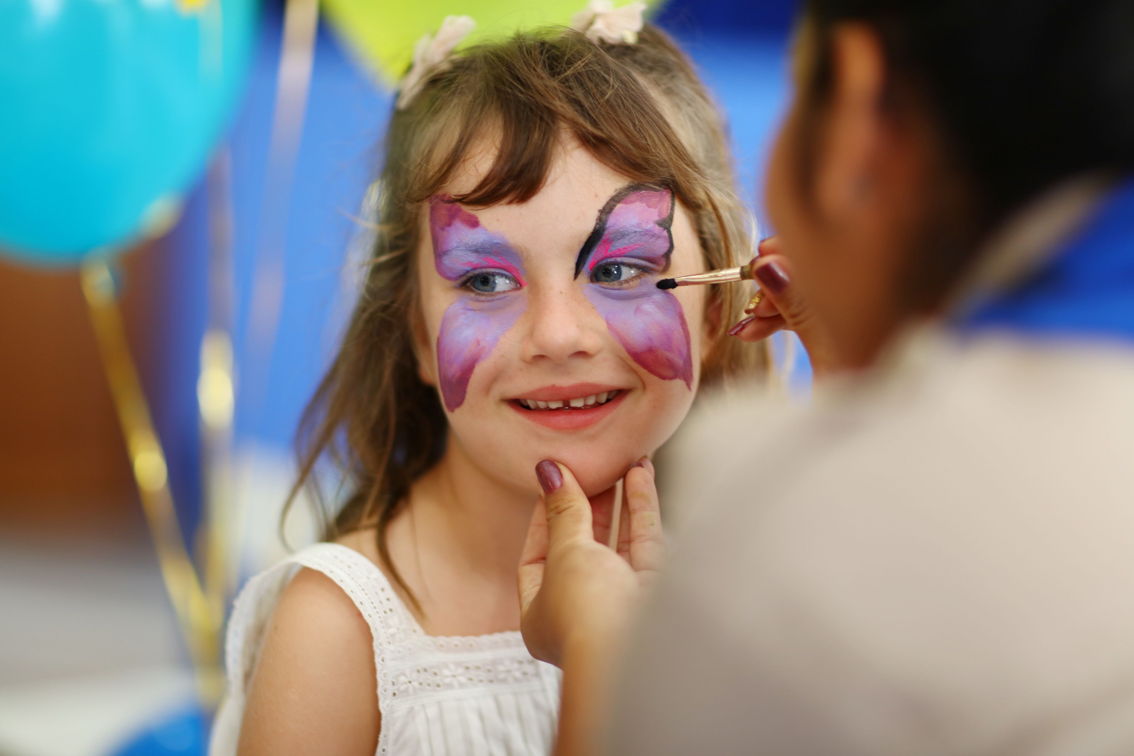 A child with face paint, a white dress, and a bow in her hair, with a person painting her face.