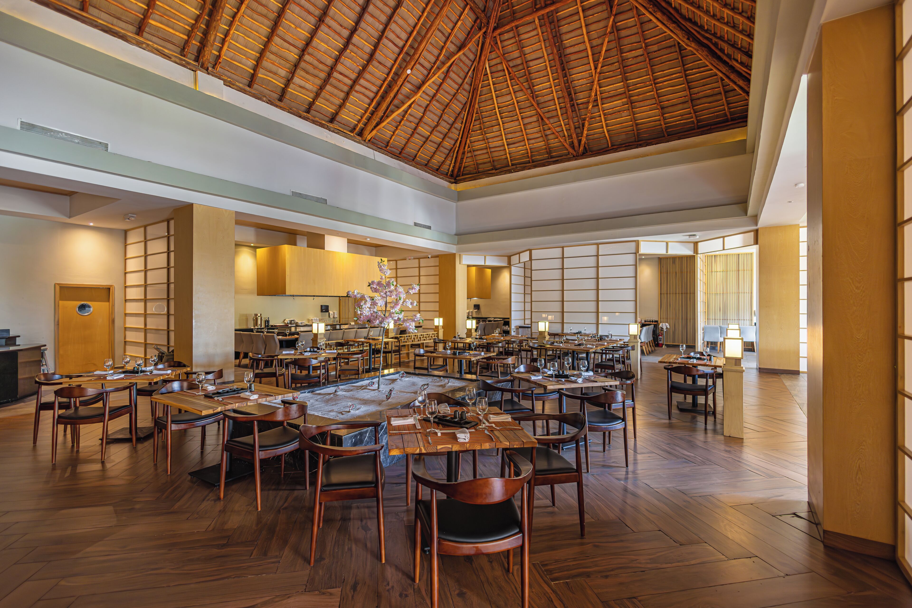 A spacious dining area with multiple tables and chairs, a high ceiling with a wooden beam structure, and a large window allowing natural light.