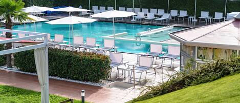 A pool area with multiple lounge chairs and umbrellas, surrounded by greenery and a building in the background.