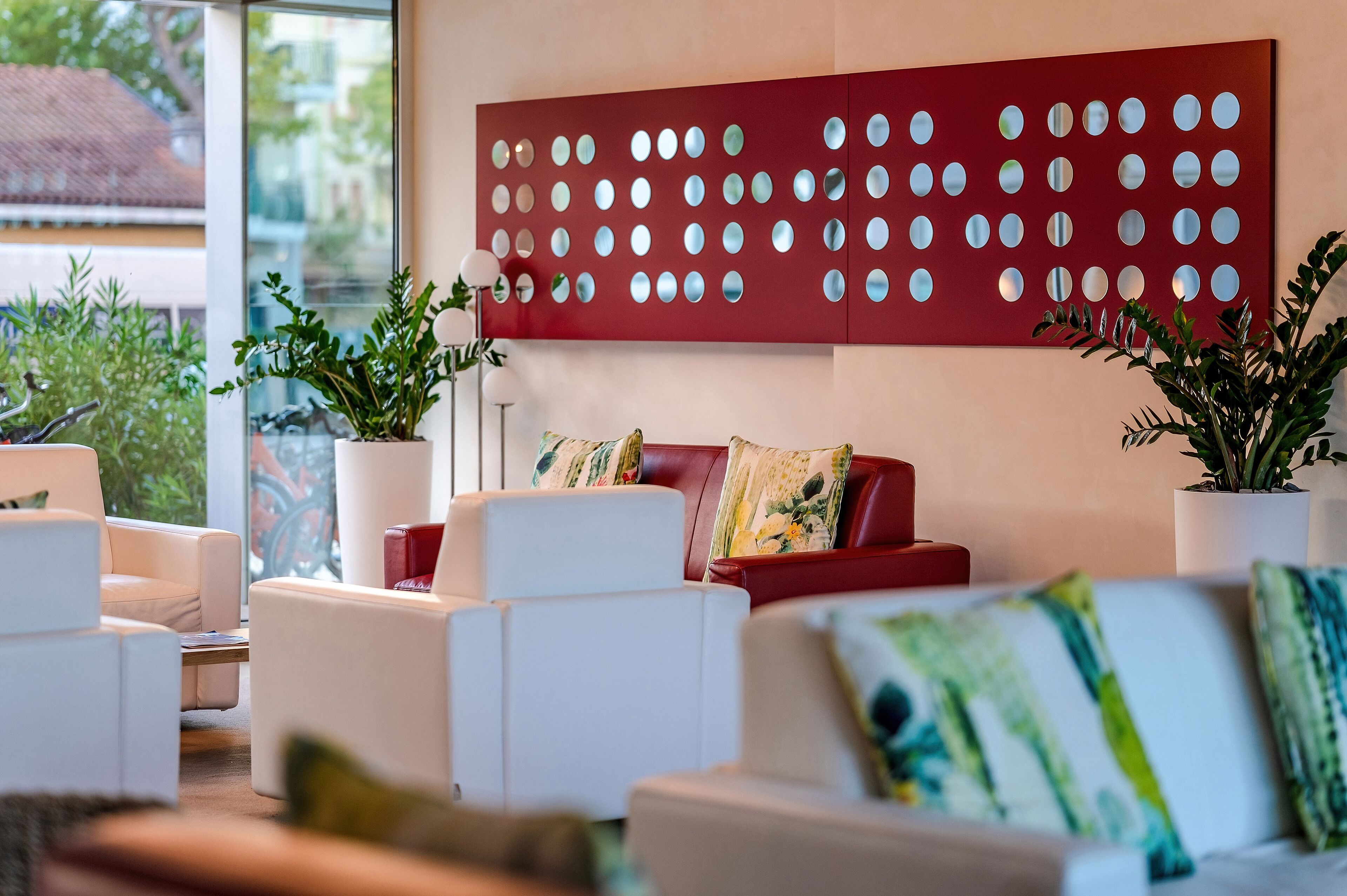 Ein modernes Wohnzimmer mit weißen und roten Sofas, einer roten Wand mit runden Spiegeln sowie Zimmerpflanzen.