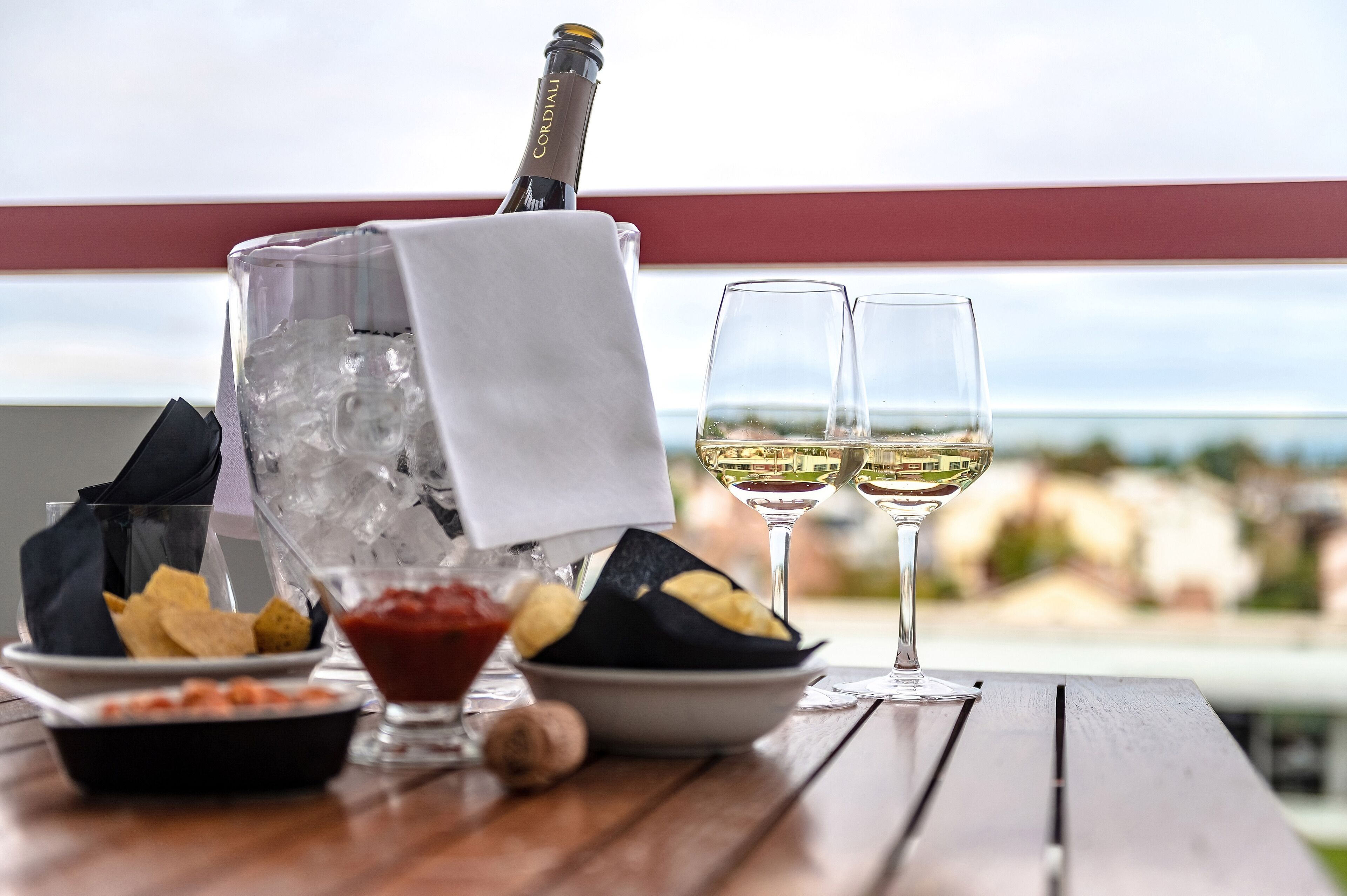 A table with a bottle of wine, two glasses of white wine, bowls of snacks, and a bottle of ketchup.