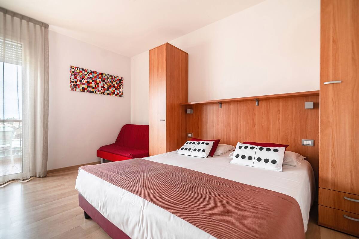 A modern hotel room with a large bed, a red chair, a wooden wardrobe, and a colorful artwork on the wall.