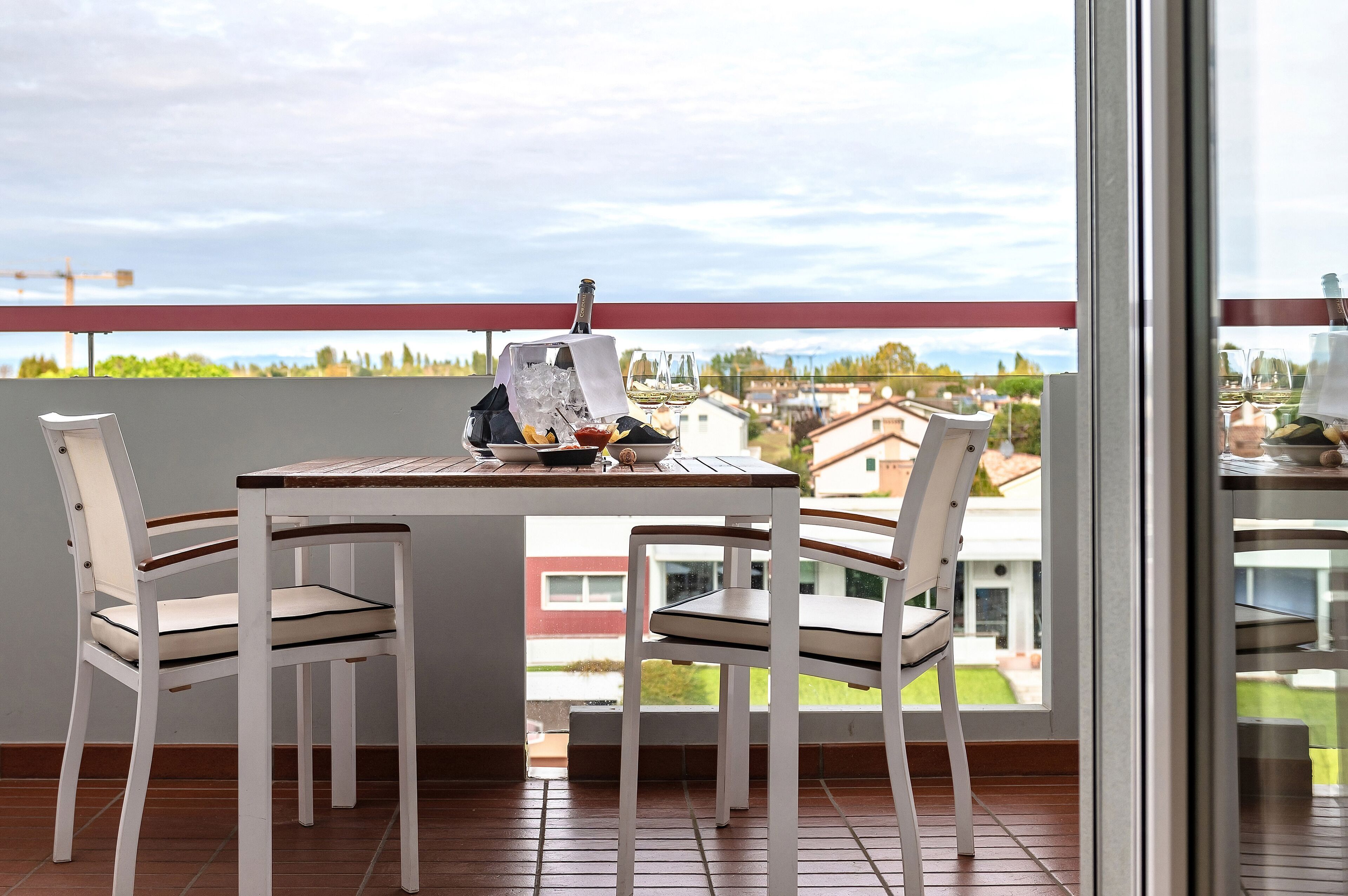 Un balcon avec une table dressée pour deux personnes, des chaises, une bouteille de champagne et une vue sur un quartier résidentiel.