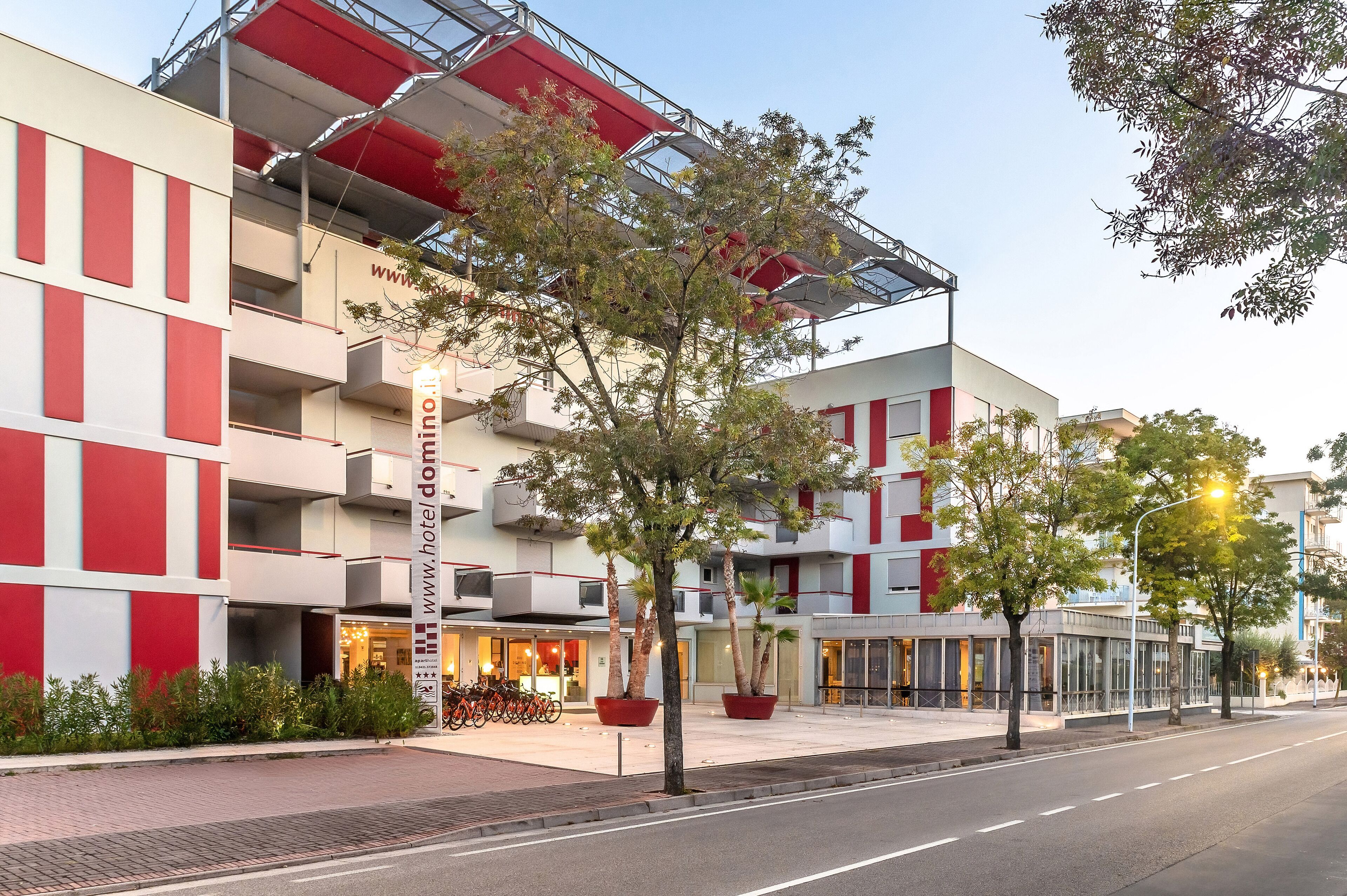 A modern hotel building with a red and white facade, multiple balconies, and potted plants in the foreground.