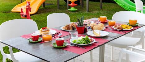 Un área de comedor al aire libre con una mesa dispuesta para el desayuno, rodeada de sillas blancas, y un parque infantil con toboganes al fondo.
