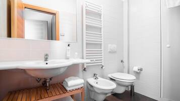 Et moderne badeværelse med vask, bidet og toilet i hvidt, et trækommode og en håndklæderække.