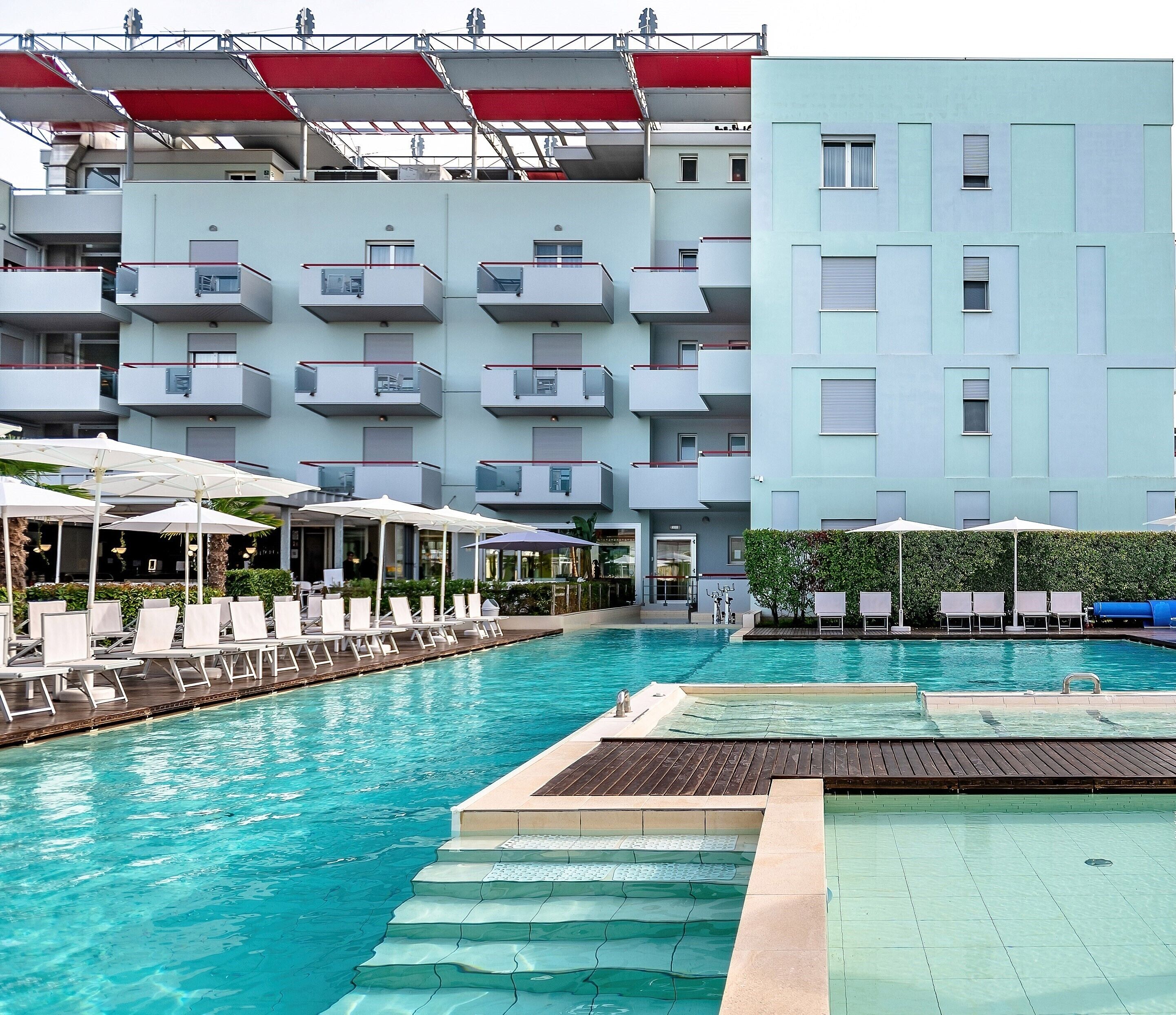 A swimming pool with sun loungers and umbrellas, surrounded by a multi-story building with balconies.
