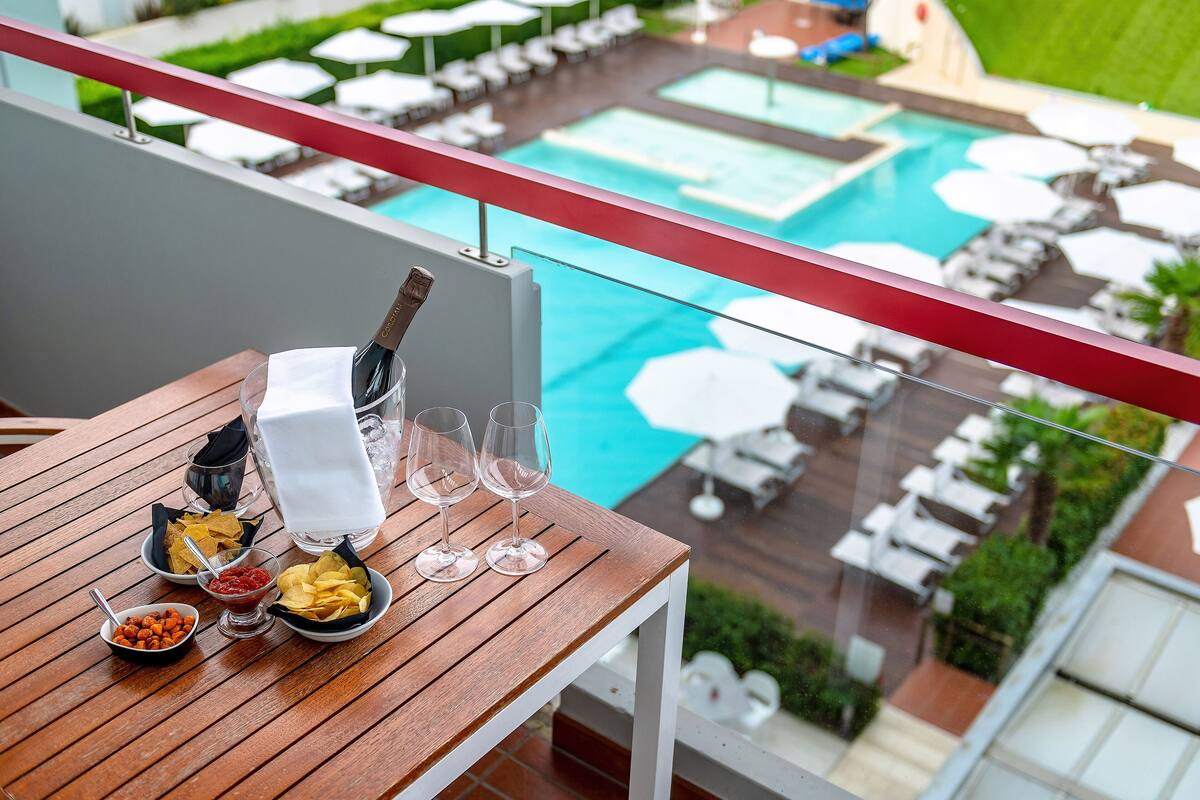 A balcony with a wooden table set for dining, overlooking a swimming pool area with lounge chairs and umbrellas.