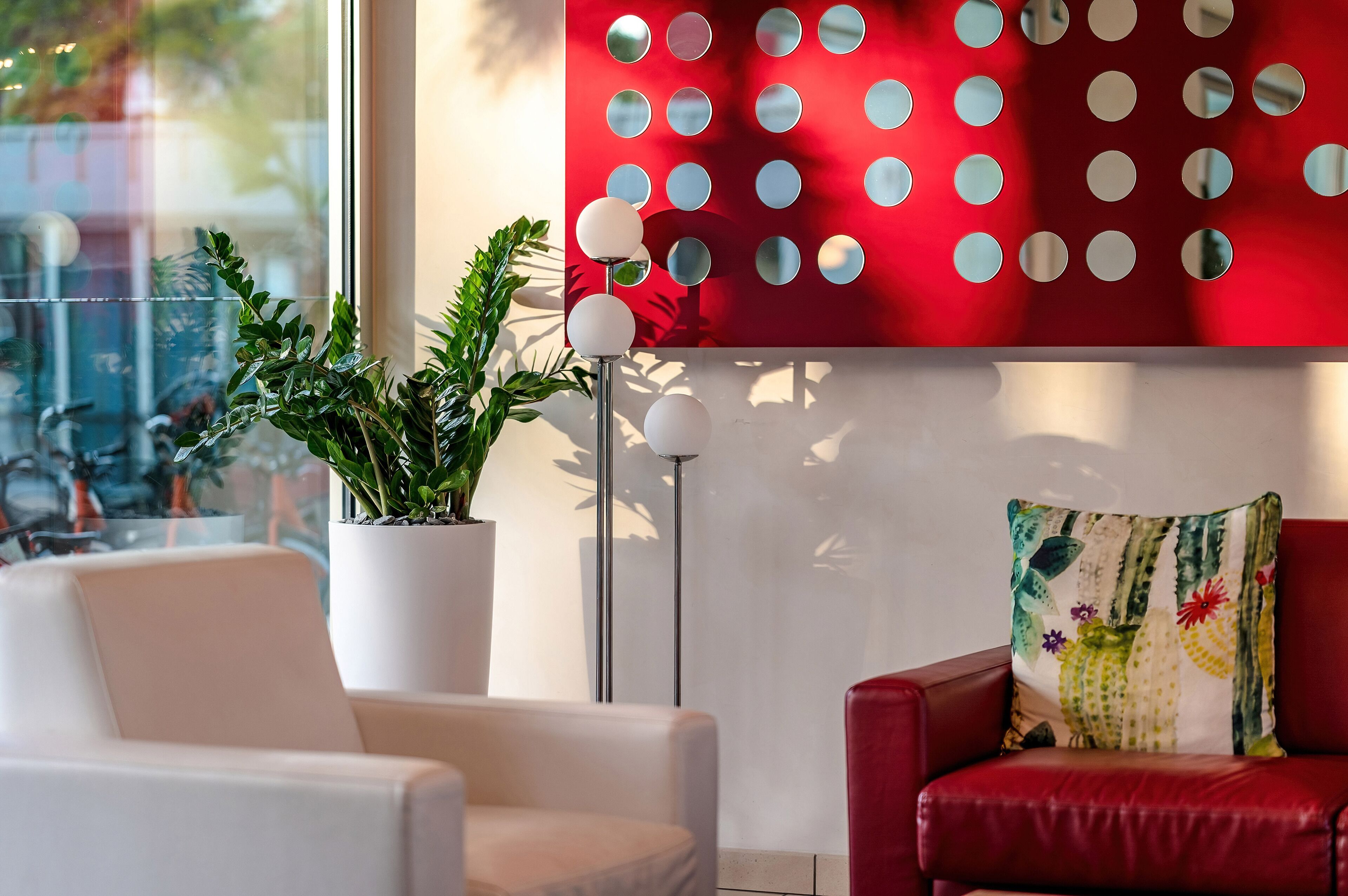 A modern room with a red wall, a white armchair, a red leather sofa, a potted plant, and a floor lamp.