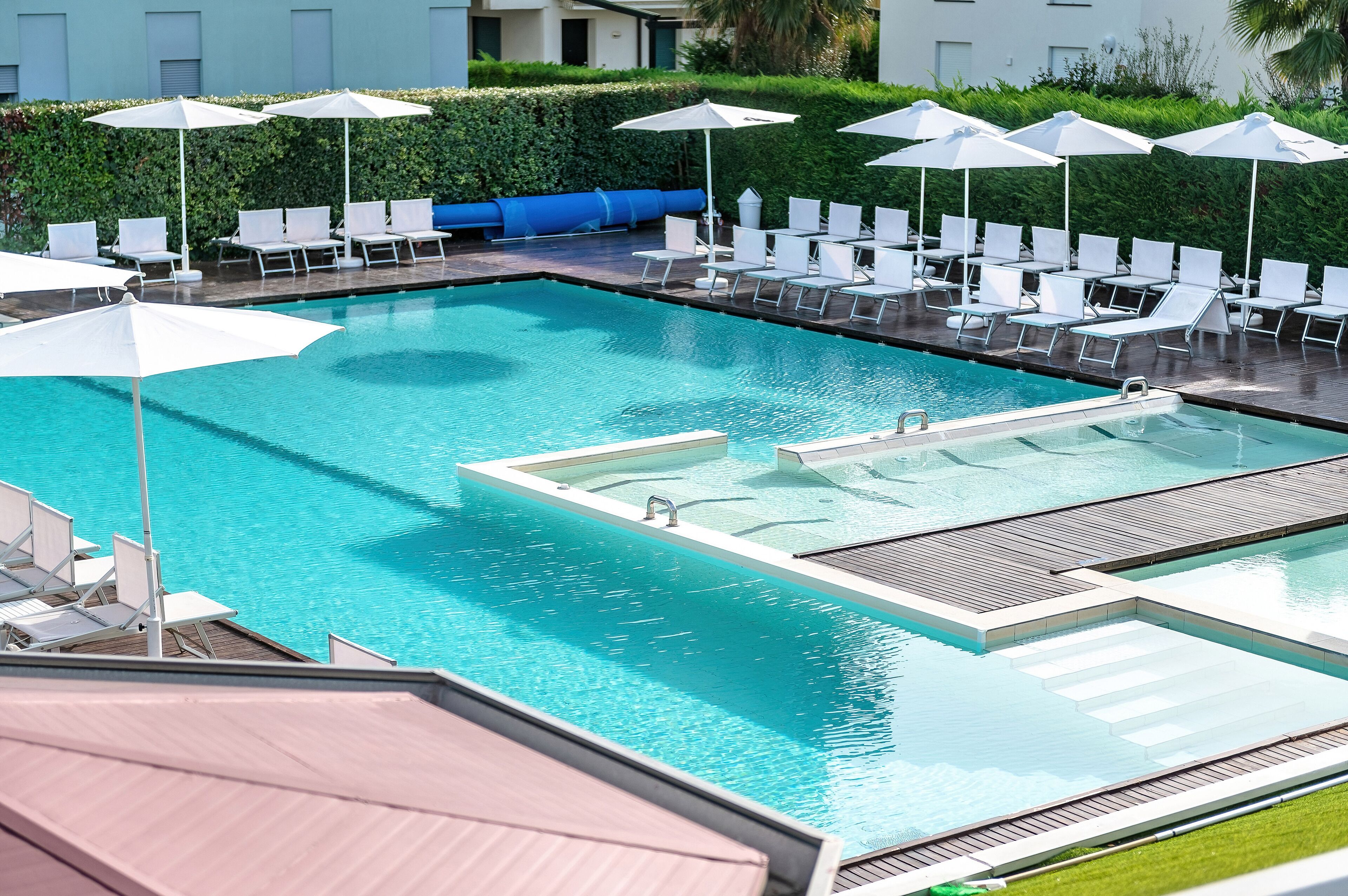 A rectangular swimming pool with a wooden deck, surrounded by white lounge chairs and umbrellas.