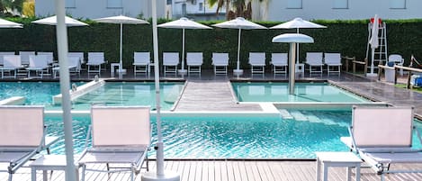 Un espace piscine doté de plusieurs chaises longues et de parasols, d’une terrasse en bois et d’une vue sur un bâtiment en arrière-plan.