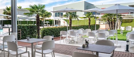 An outdoor patio with white tables and chairs, umbrellas, and palm trees.