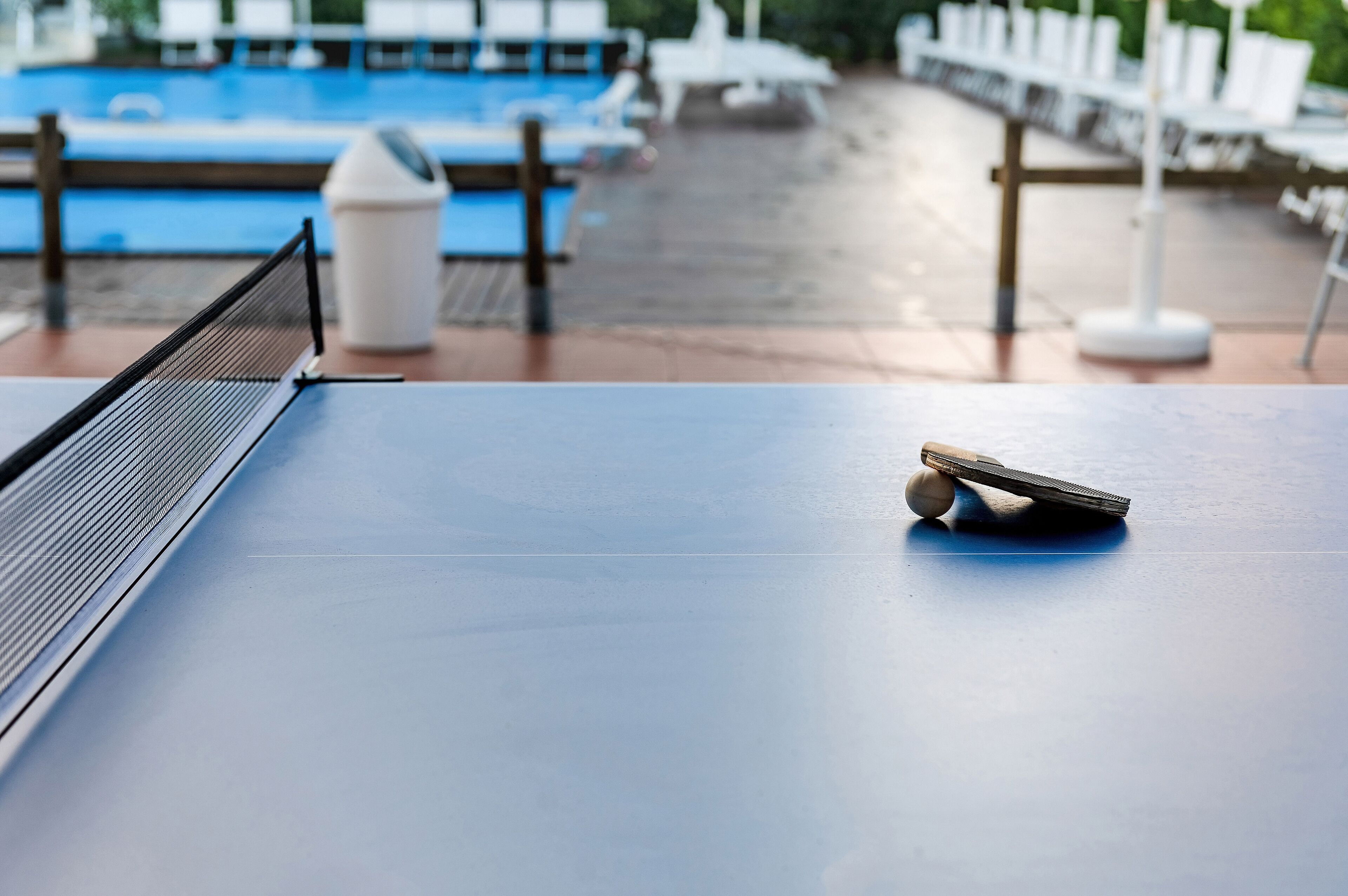 A ping pong table with a ball and paddle, a trash can, and a pool in the background.