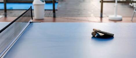 Taula de ping-pong amb una pilota i una paleta, un safataire i una piscina al fons.