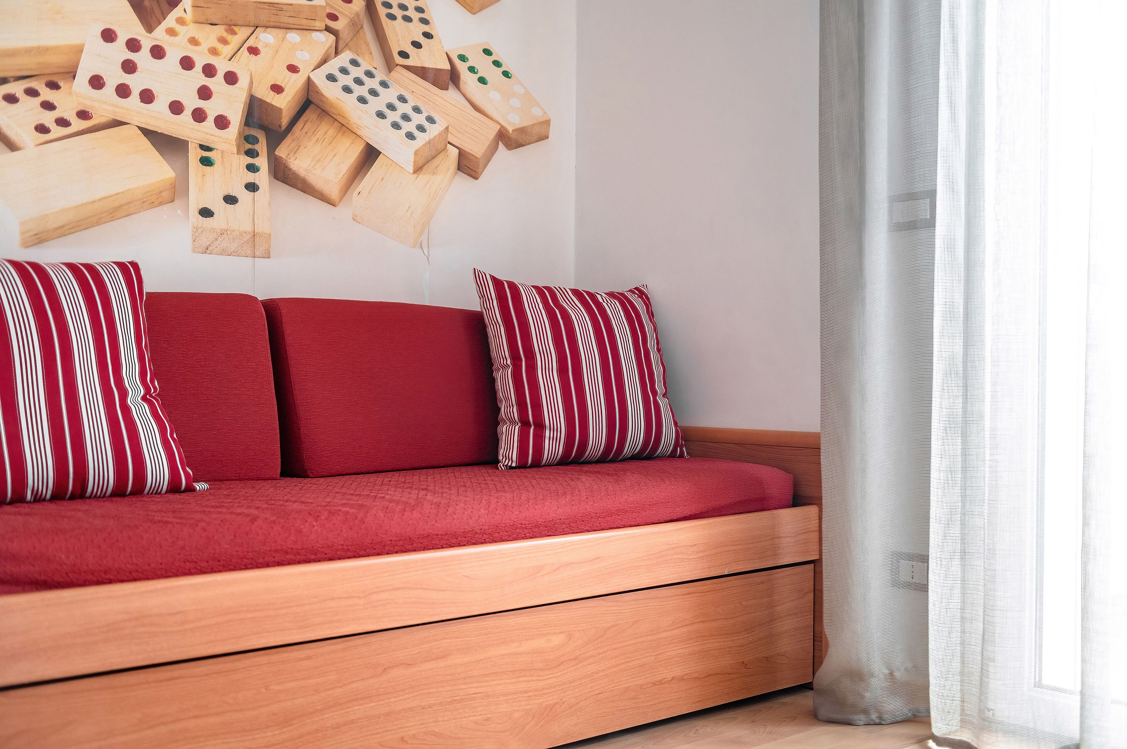 A wooden sofa with red cushions and striped pillows, a wall with wooden dominoes, and a sheer white curtain.