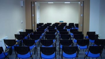 A conference room with rows of black chairs with blue cushions, a podium at the front, and a plain white wall.