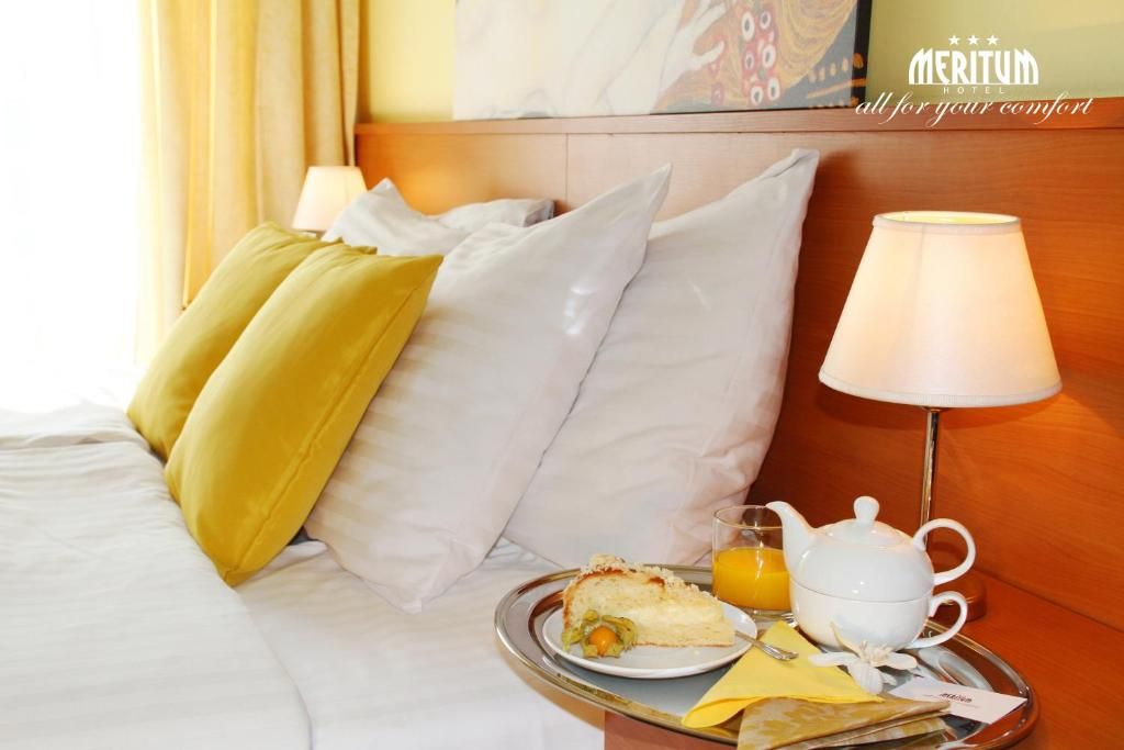 Una habitación de hotel con una cama, almohadas, una bandeja con alimentos para el desayuno y una lámpara.