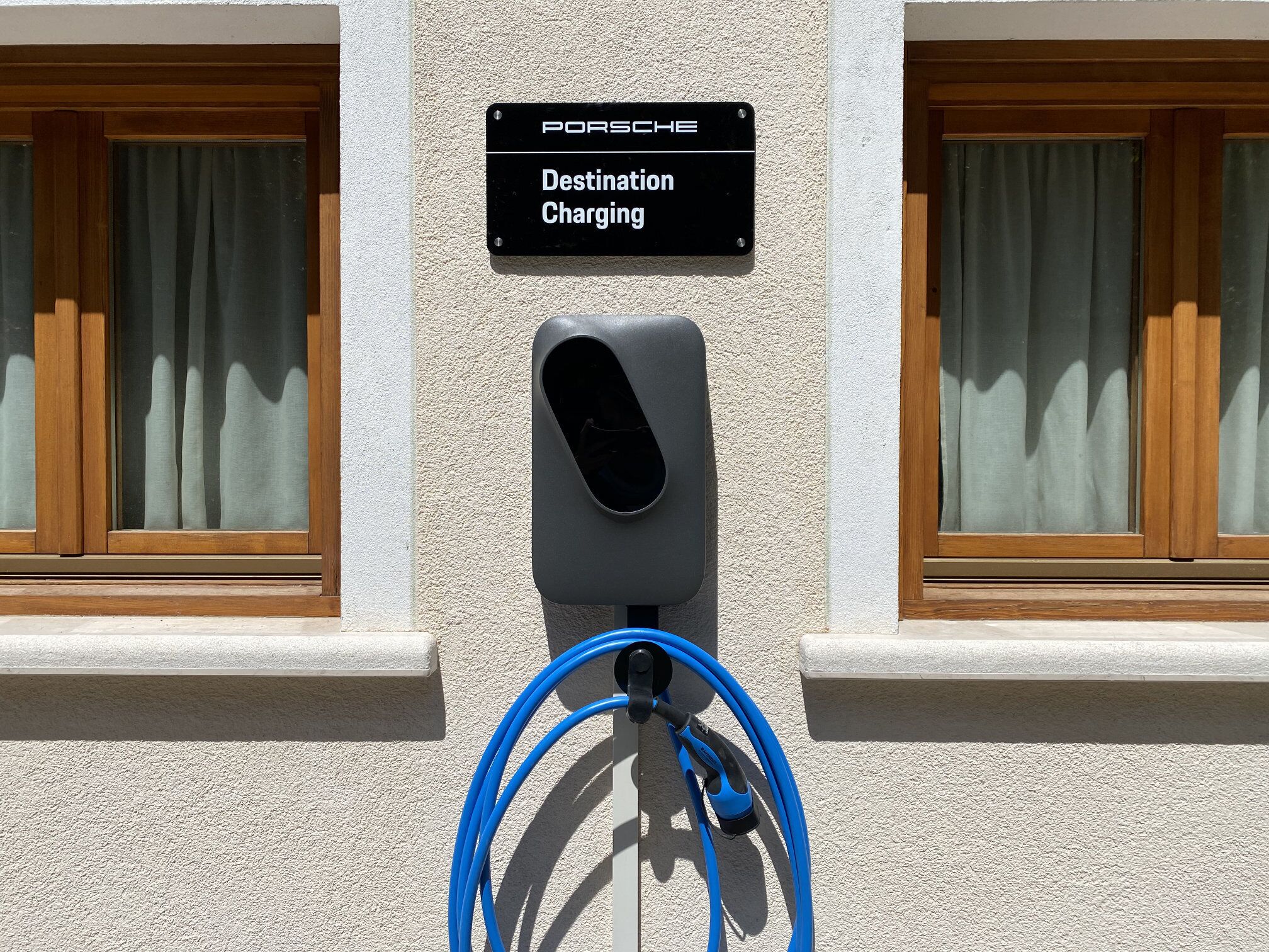 A destination charging station for Porsche vehicles, mounted on a building with wooden window frames and a textured wall.