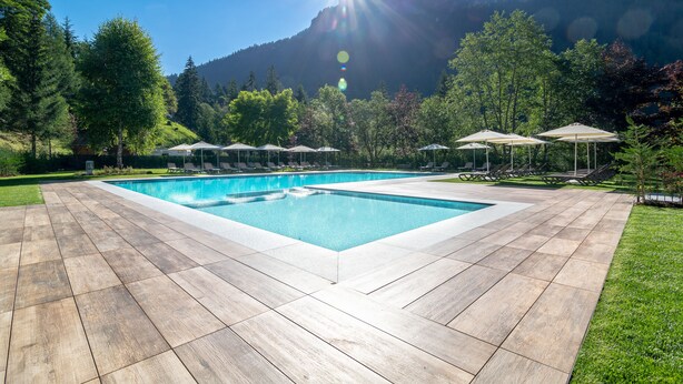 A rectangular swimming pool with a wooden deck, surrounded by lush greenery and mountains in the background. - Hotel La Maison Wellness & SPA (Alleghe)
