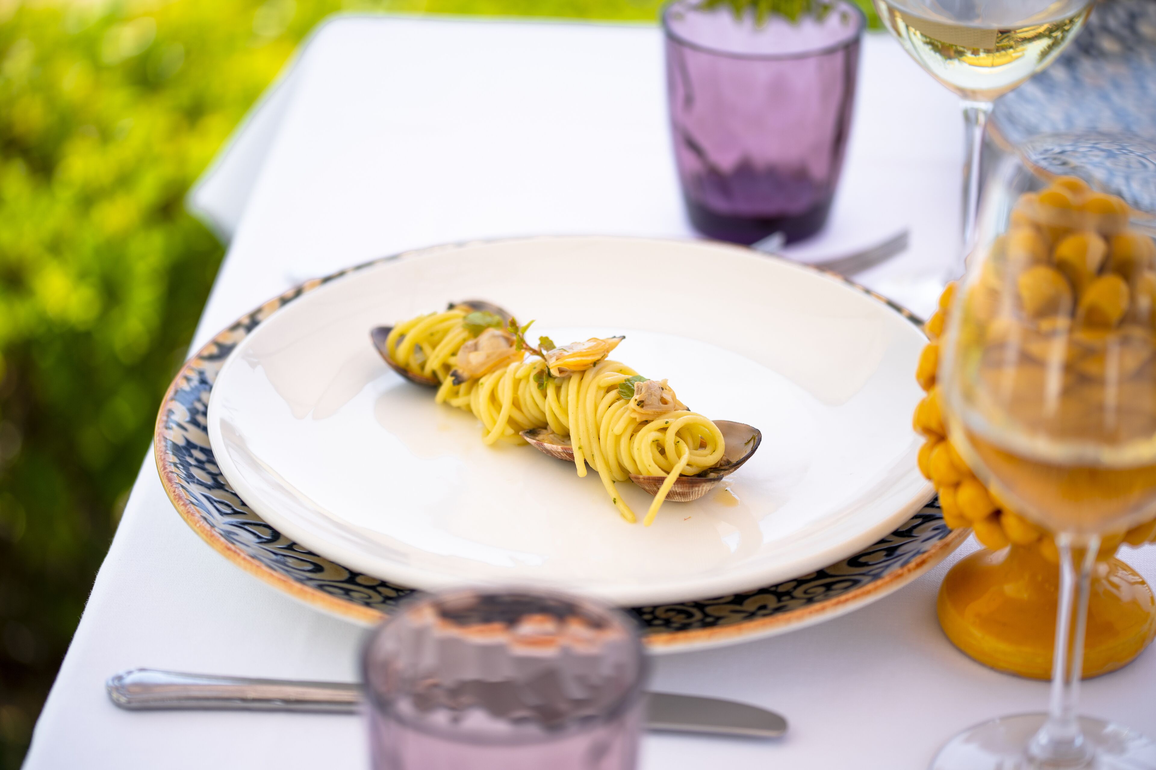 A dining table with a plate of spaghetti, a glass of white wine, and a glass of yellow beverage.