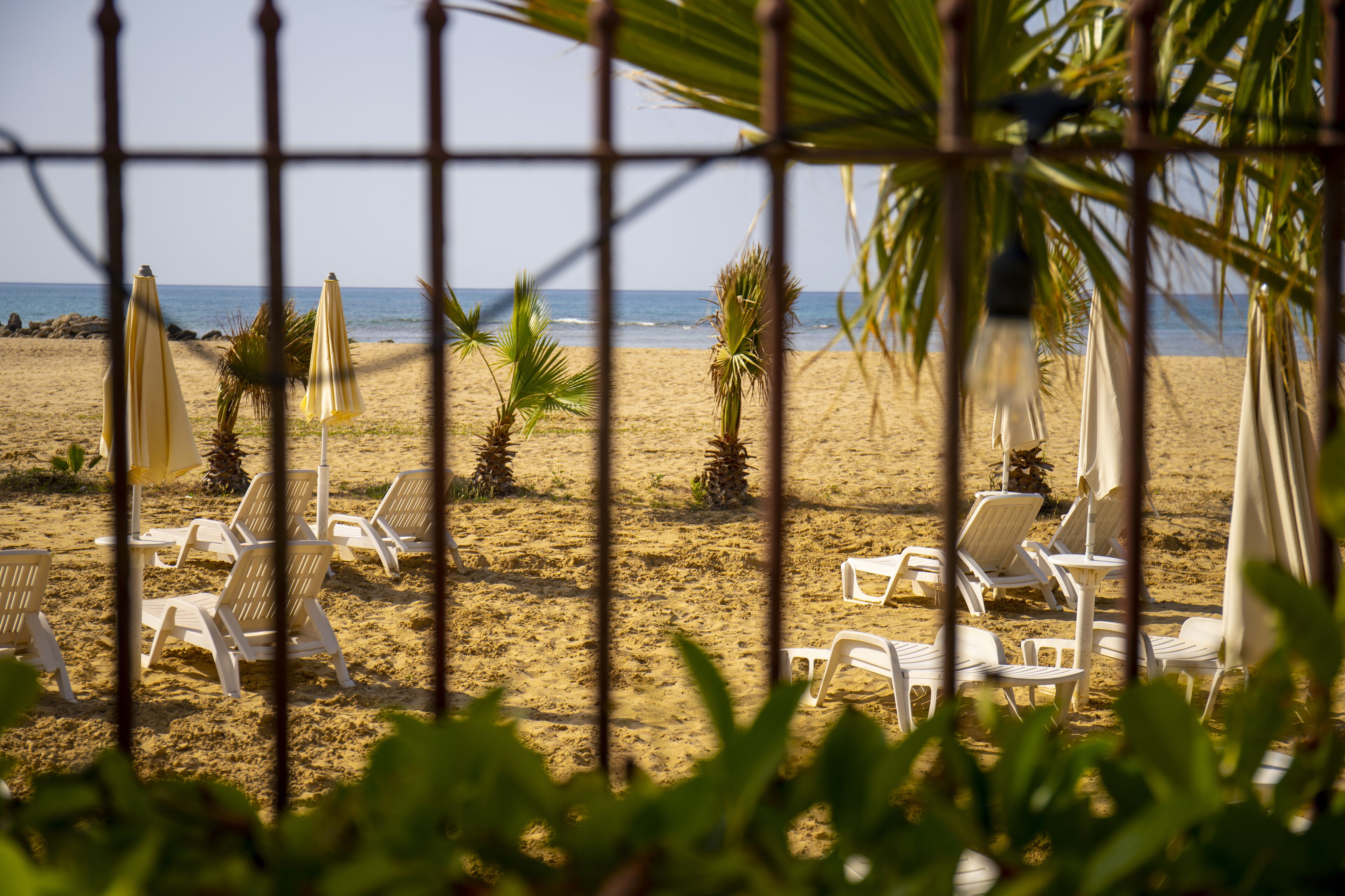 Een strand met witte ligbedden en gele parasols, zichtbaar door een metalen hek.