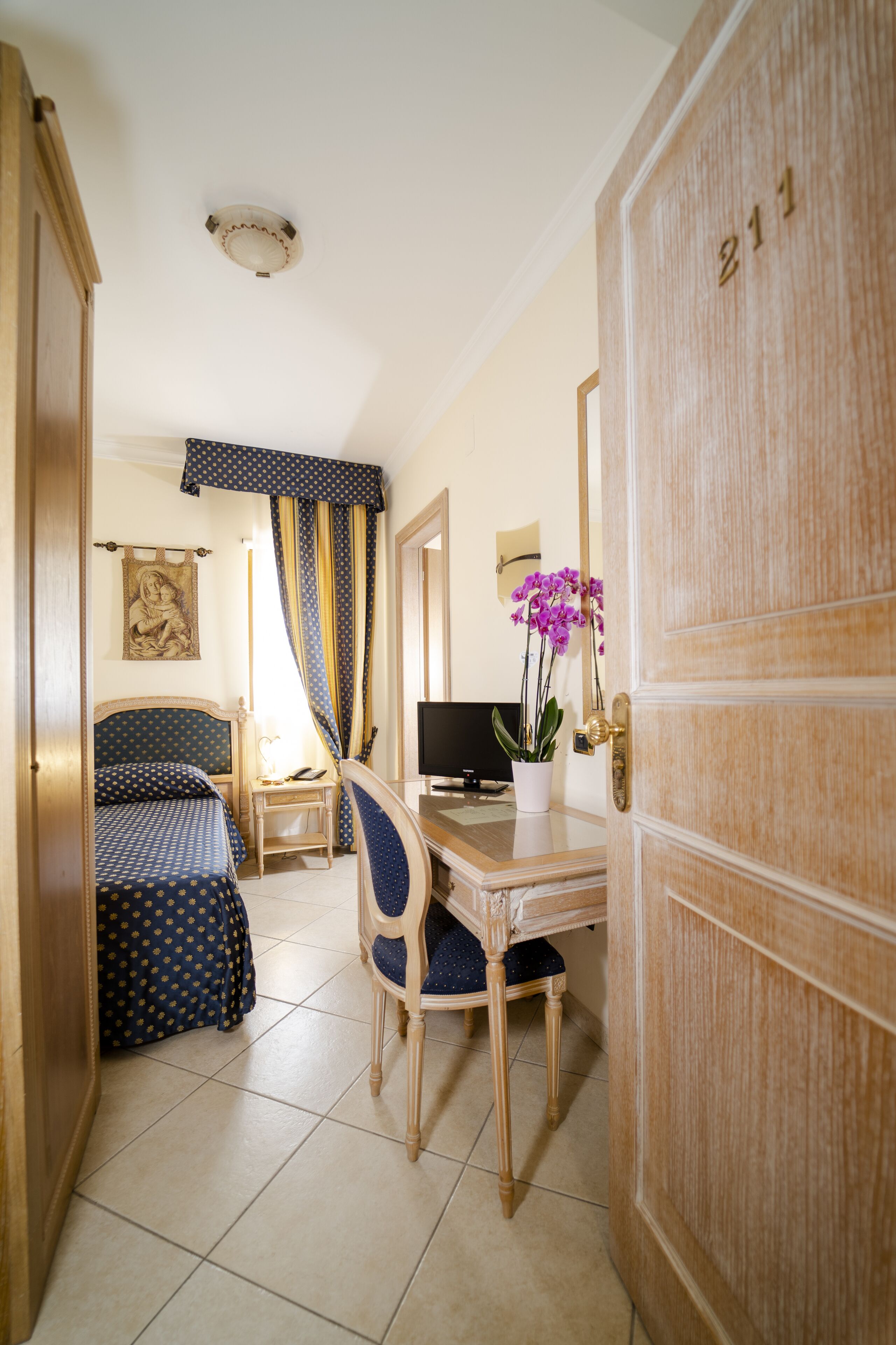 Ein Hotelzimmer mit einem Bett, einem Schreibtisch mit Computer, einem Sessel, einer Zimmerpflanze und einer Tür mit einer Nummer.