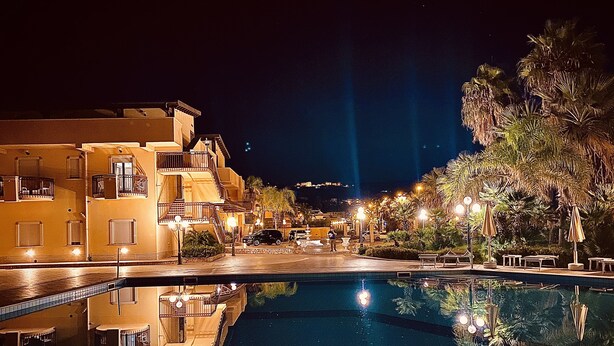 A nighttime view of a hotel with a swimming pool, reflecting the buildings and street lights.