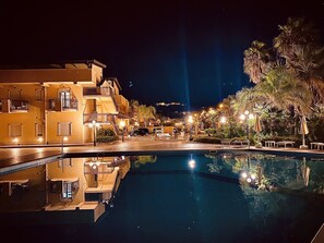 Vista noturna de um hotel com piscina, refletindo edifícios e luzes da rua.