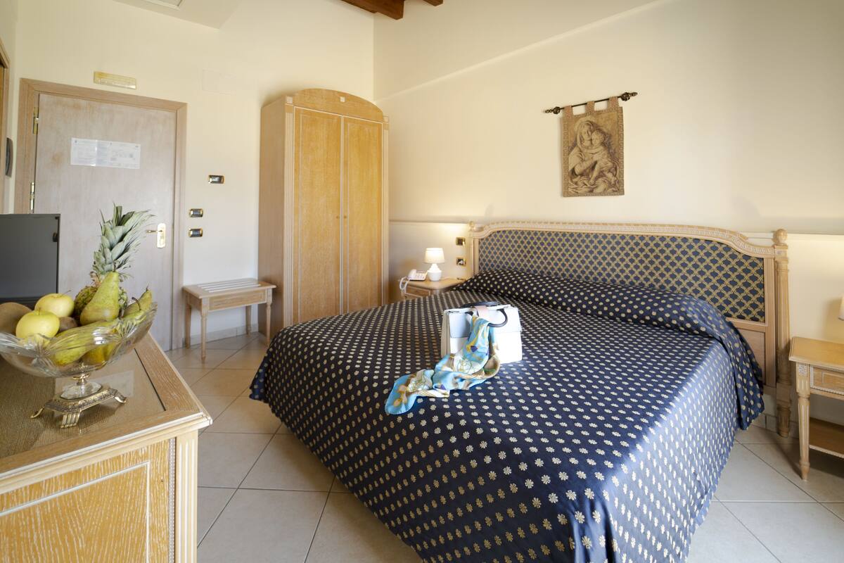 Une chambre d’hôtel avec un lit, une armoire en bois, un bureau avec un panier de fruits et une petite table avec une lampe.